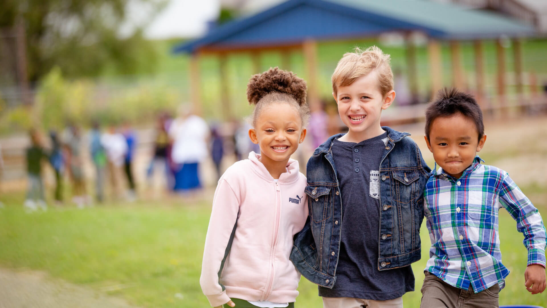 3 elementary students joined in a group pose outside at a pavilion
